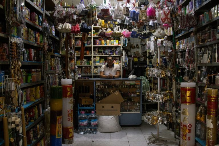 Comerciante atrás do balcão de uma pequena loja repleta de produtos variados.