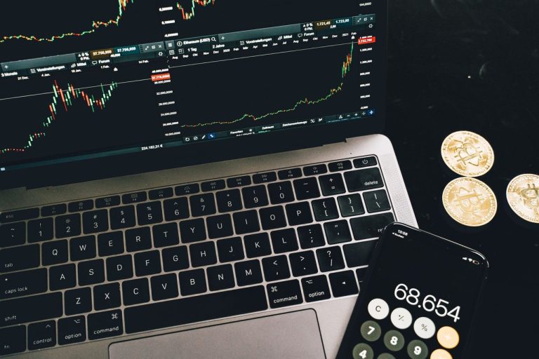 Notebook com gráficos de negociação, smartphone com calculadora e moedas de bitcoin sobre uma mesa.